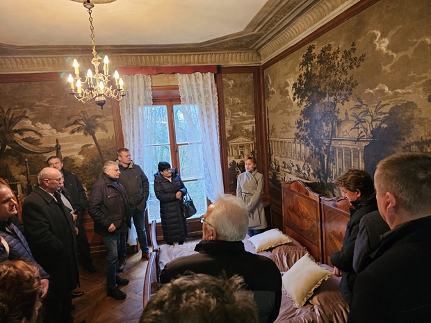 Osoby stoją w pomieszczeniu pałacu w Walewicach wytapetowanym w motywy przyrodnicze w dominującym kolorze brunatnym. W pomieszczeniu widoczny zabytkowy żyrandol, dwa łóżka i stare firany. Na zdjęciu widać ponad 10 osób.